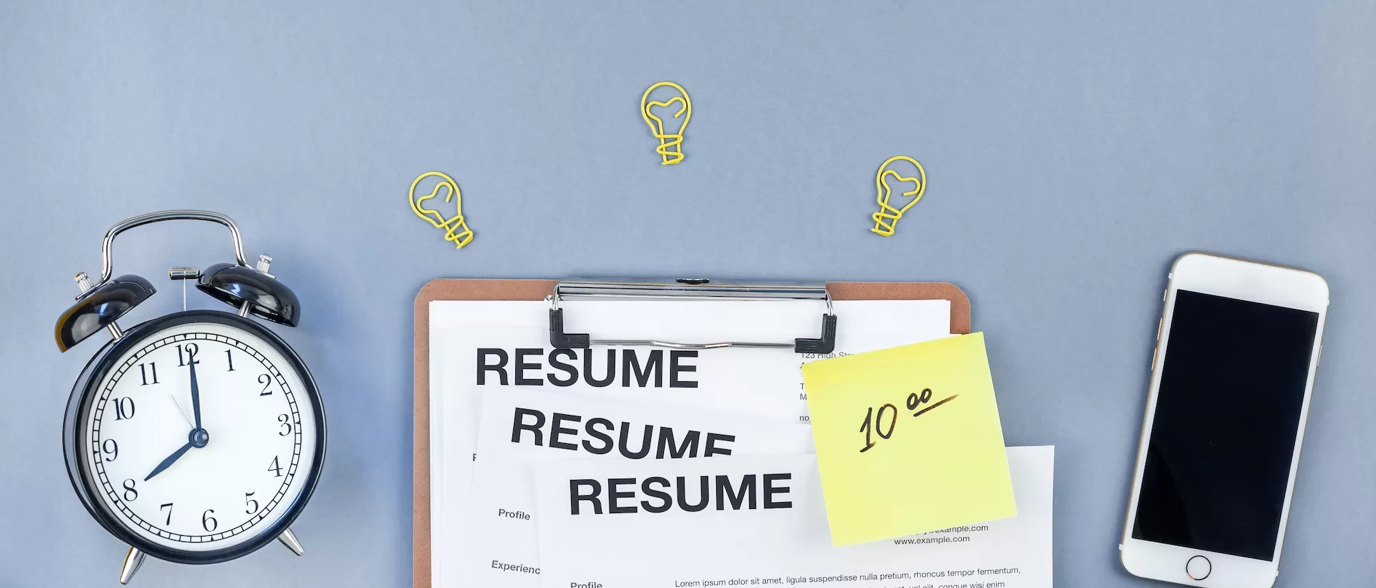A clipboard holding resume pages next to a sticky note and a phone on a desk